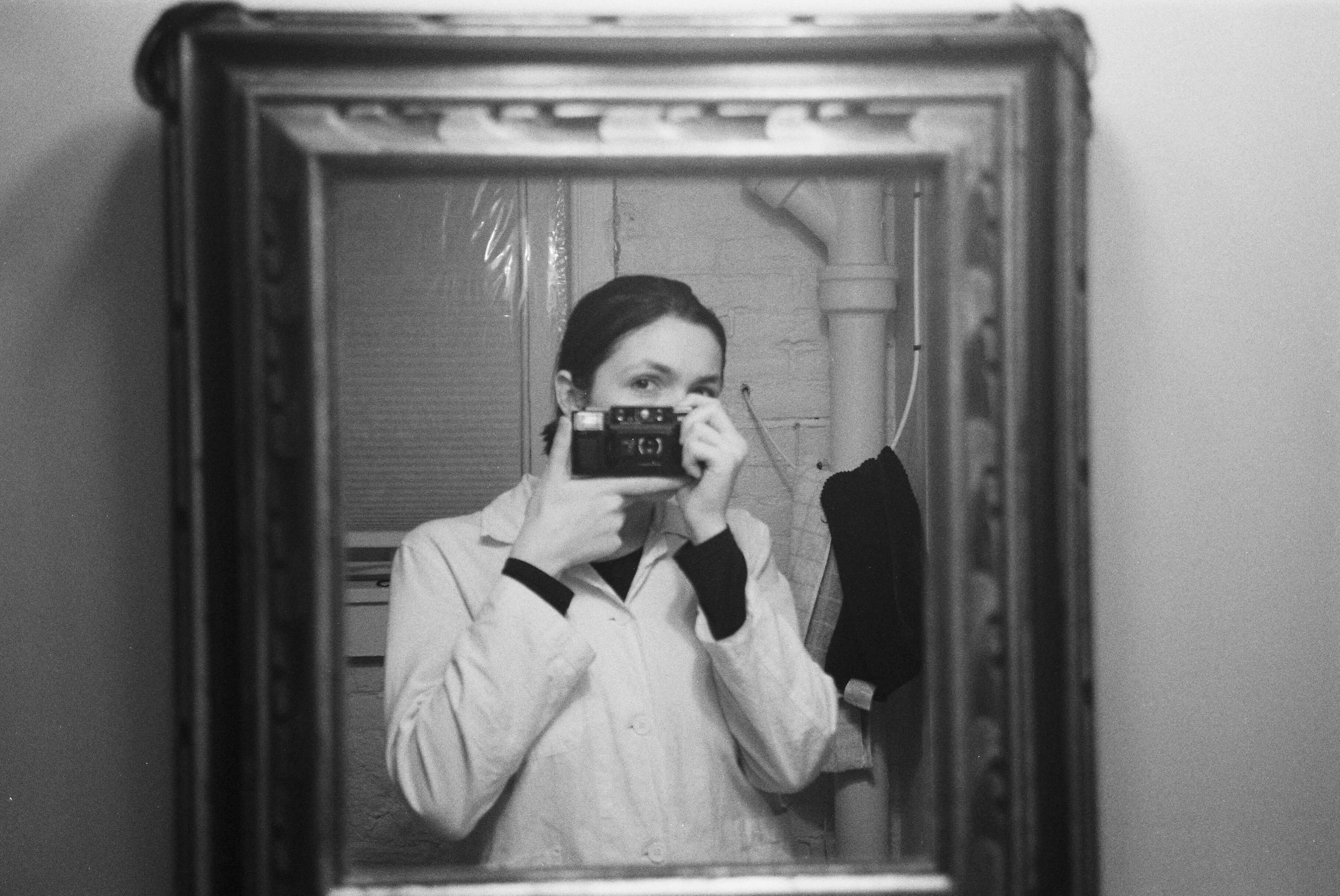 a black and white film photo from early 2020, i face my late aunt rosemary's mirror, holding a film camera that was gifted to my father from my grandmother for his graduation in the '80s. the camera covers most of my face, only one eye is fully visible. my hair is back. my white button up coat resembles a lab coat. i have a black long sleeve shirt under.
      i am in the bathroom of my basement apartment, i am the nanny to the owners of the house. it was formerly F.Scott Fitzgeralds home, while living there (1933-34) his friend wrote in his journal: “The case of F. Scott Fitzgerald has become distressing. He is a boozing in a wild manner and has become a nuisance.”